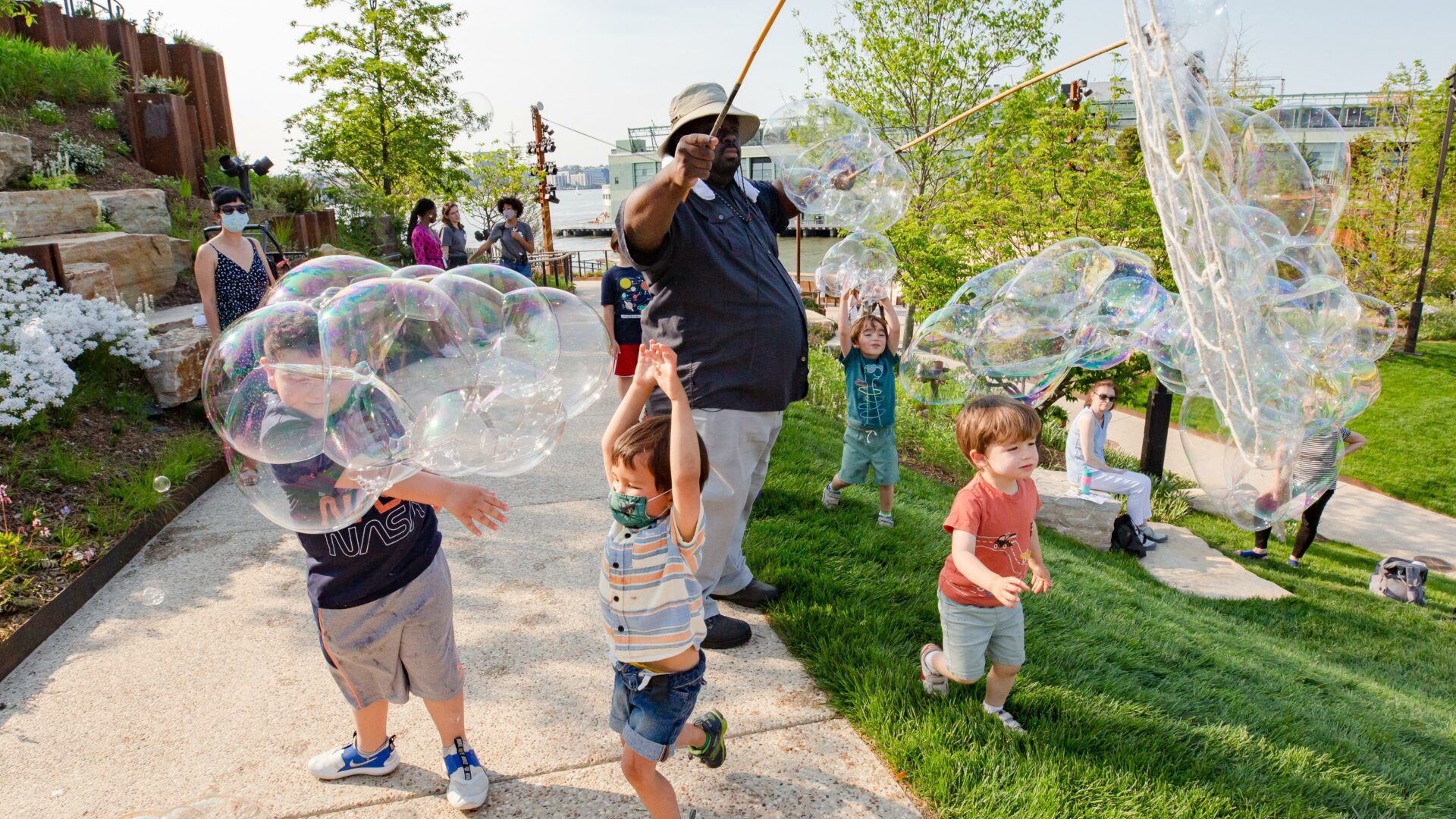 A man creates large bubbles while children frolick on the lawns