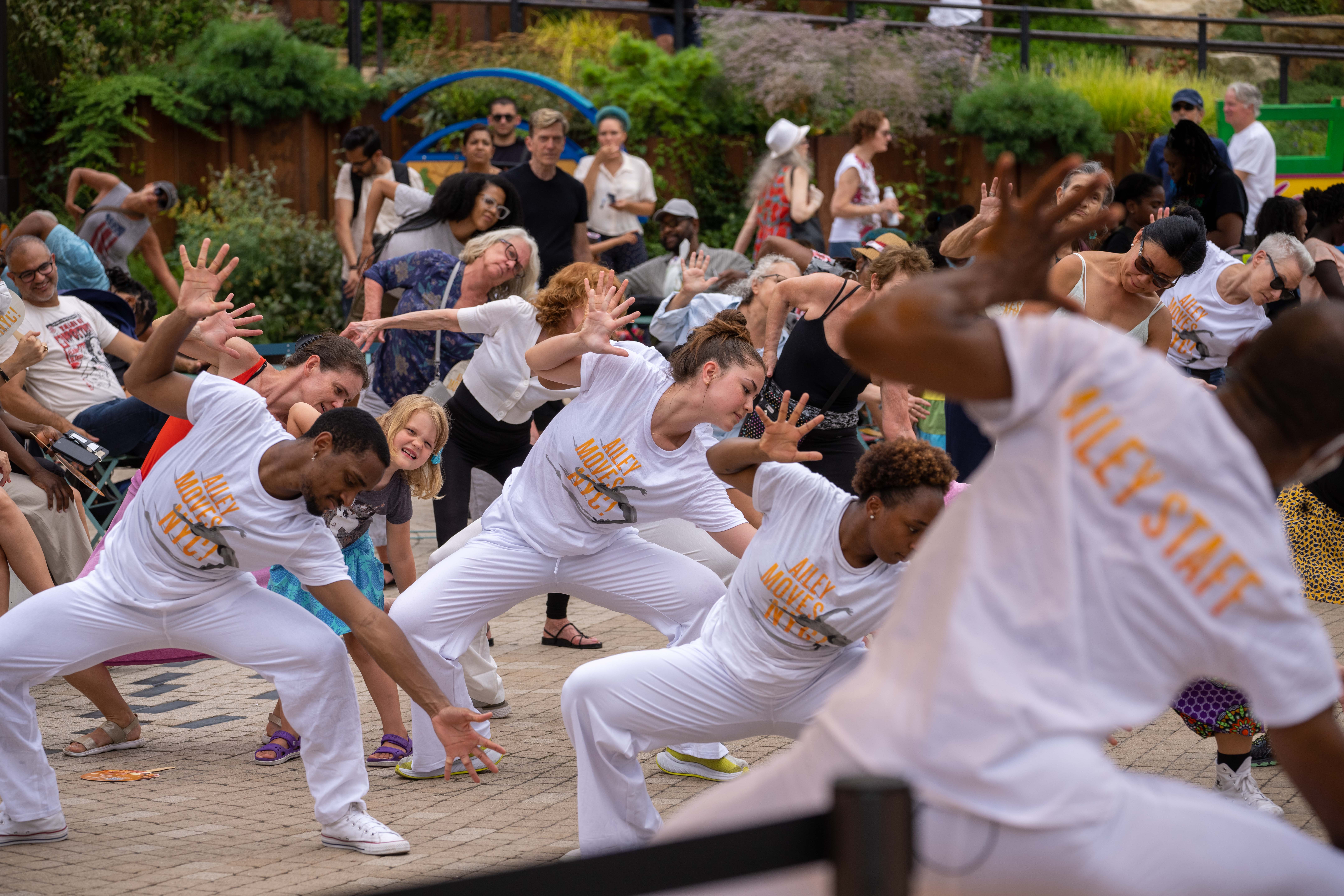 A group of dancers lean to their right showing park visitors how to do a dance move in front of lush greenery.