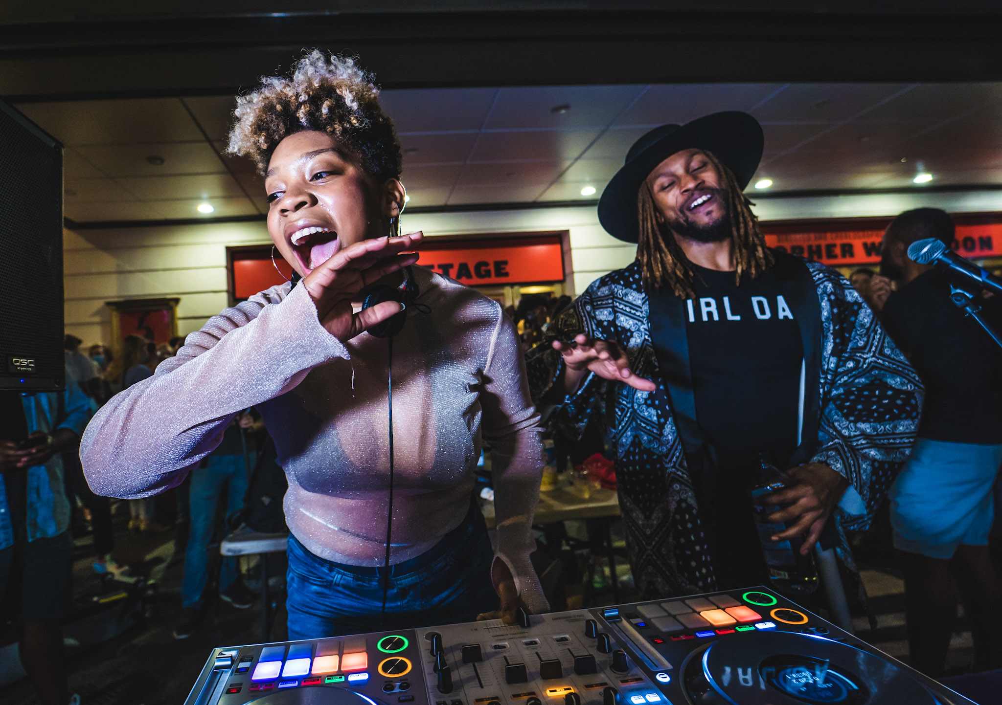 Two people joyously stand behind a DJ booth dancing.