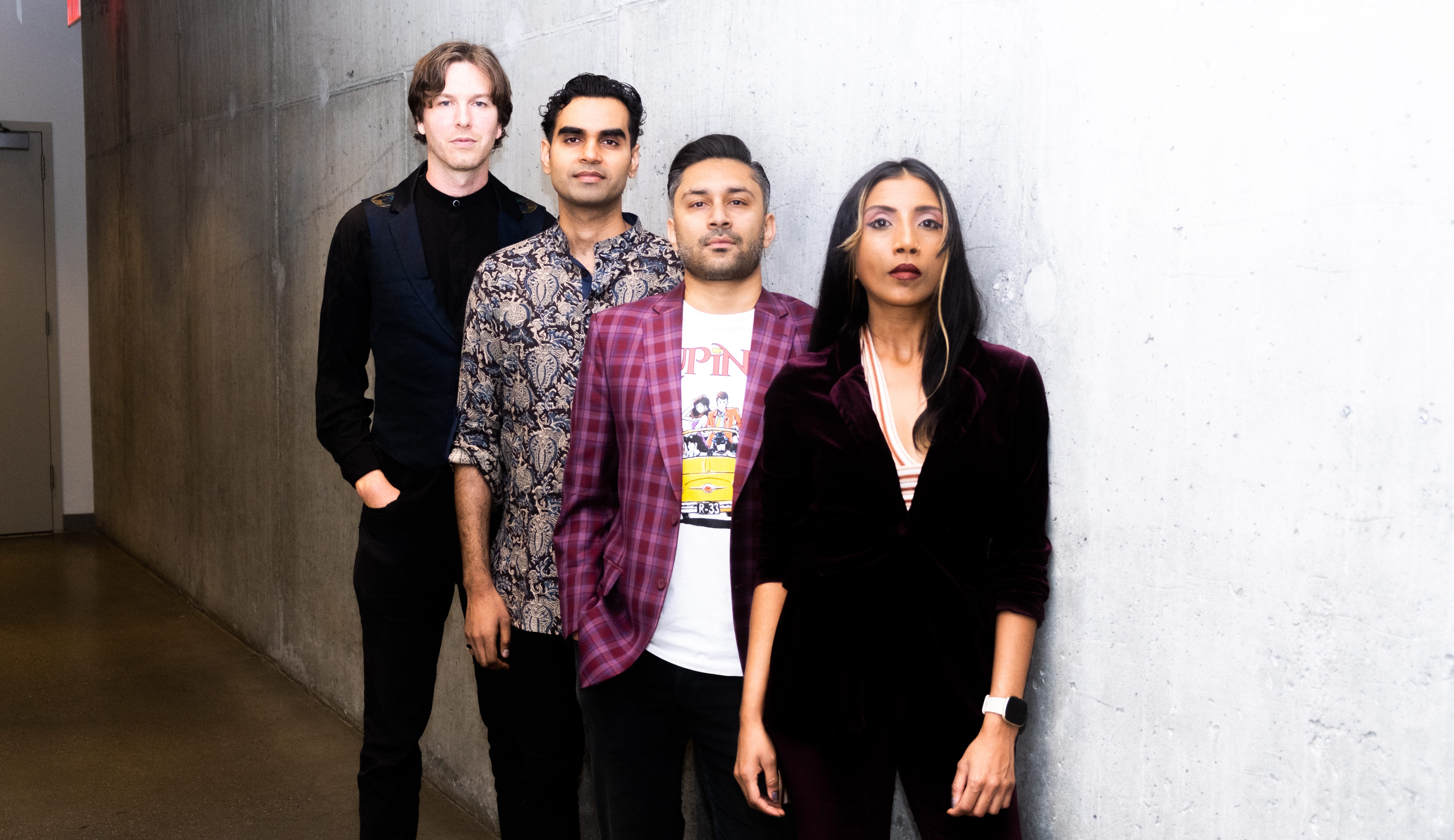 A group of people stand in a row from shortest to tallest against a plain off-shite wall.