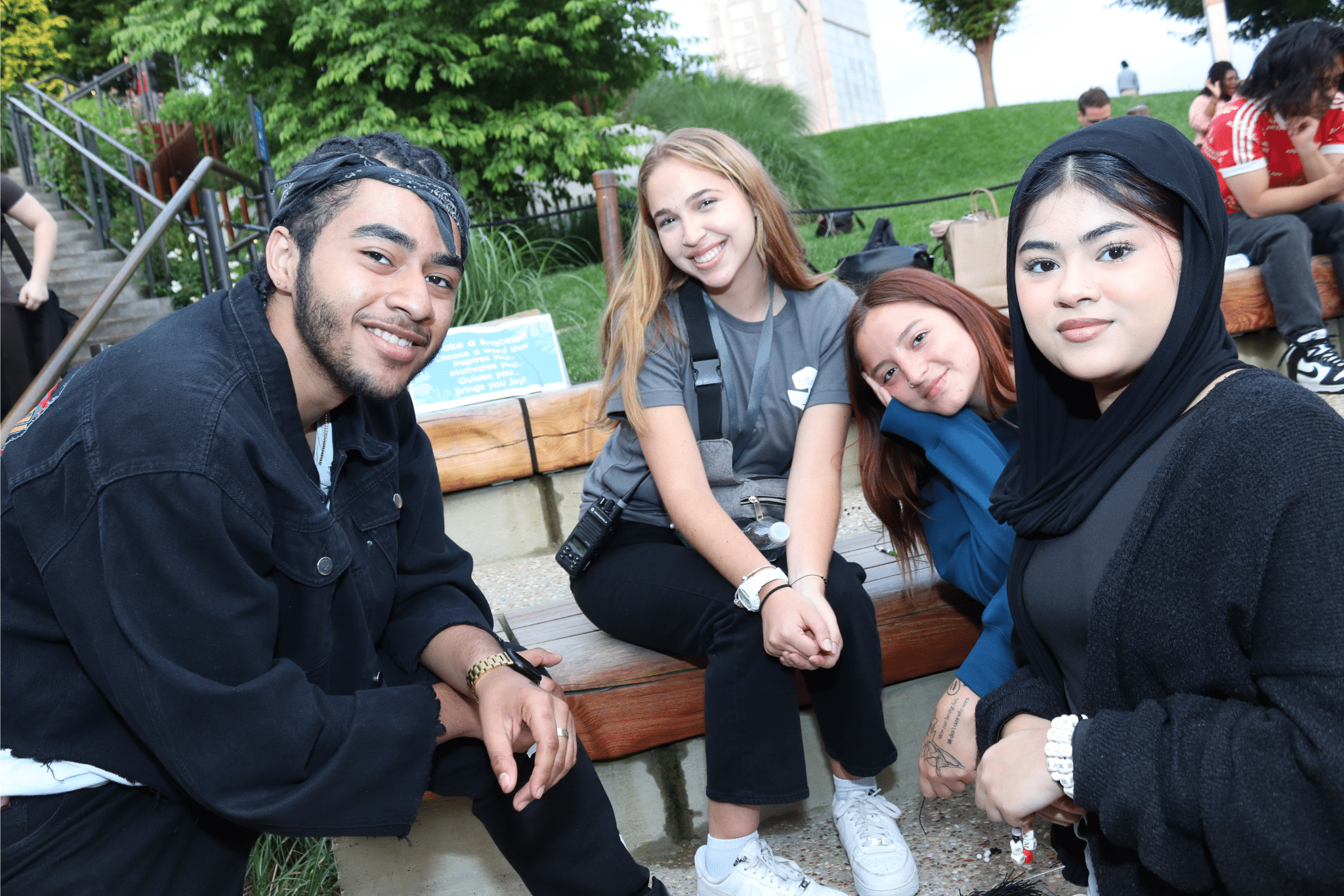 Four smiling teenagers of different races sit on a bench in a green park.