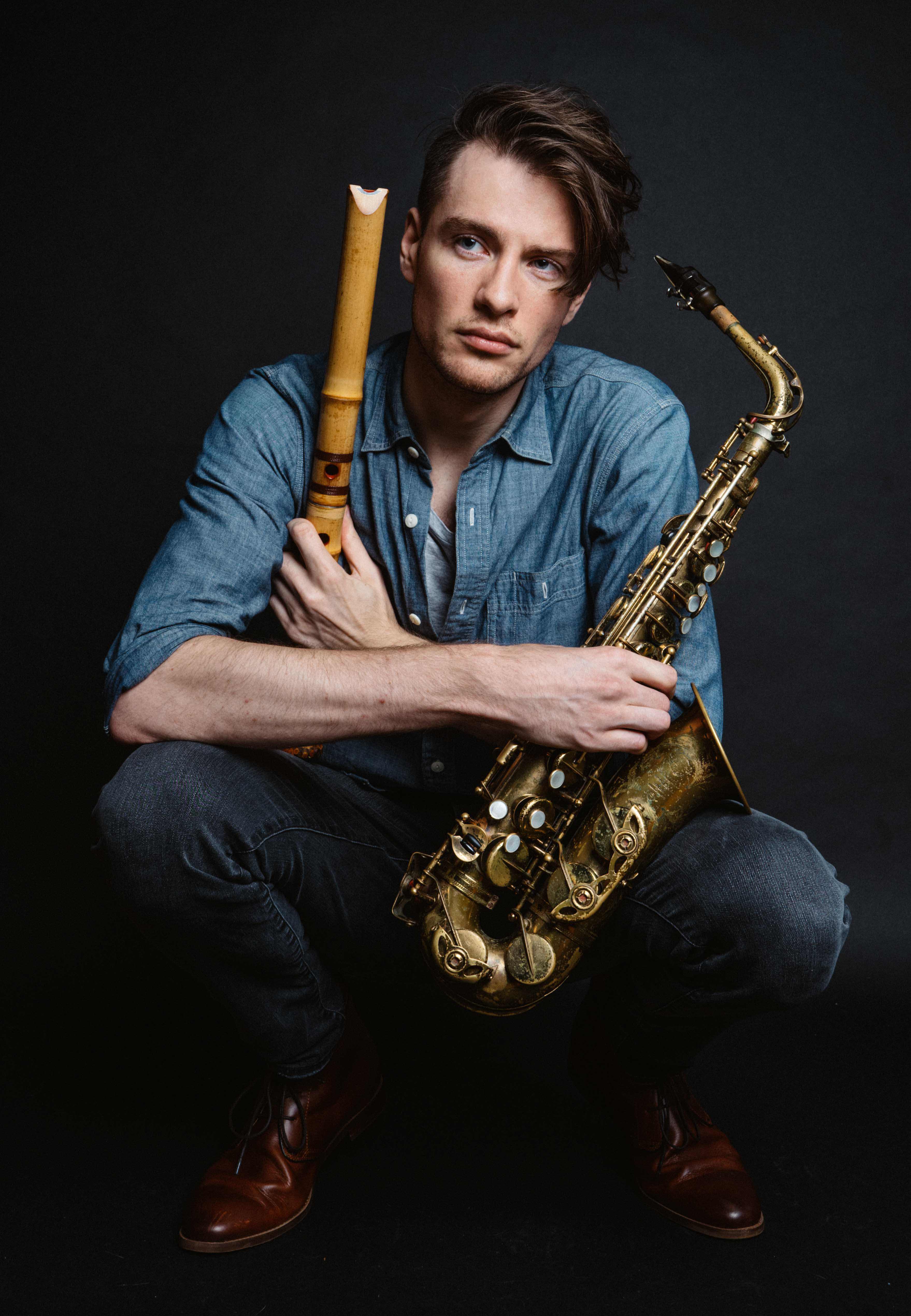 A person kneels down holding a saxophone. He is wearing a long sleeve blue button down shirt.