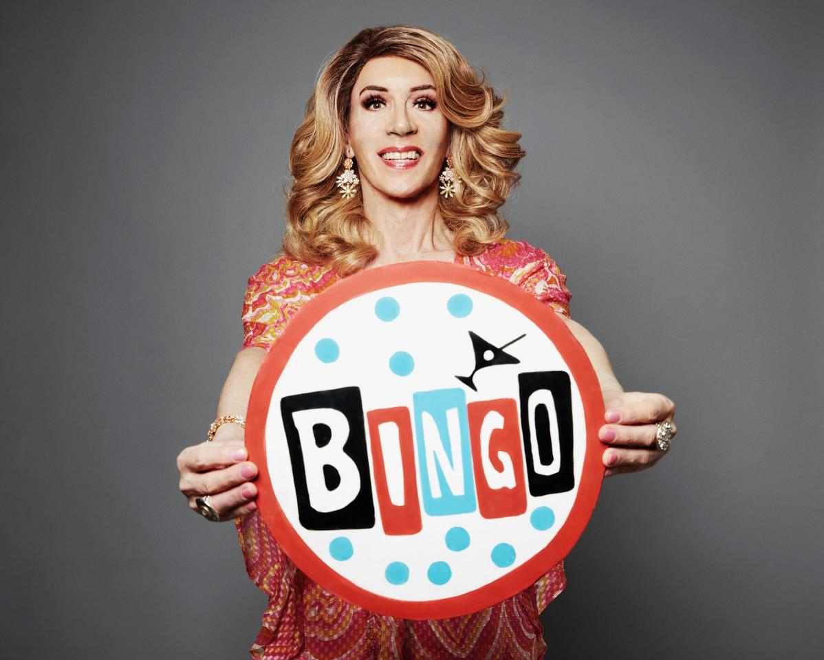 A person with wavy blonde hair and a red outfit holds a sign in front of them that says Bingo with a martini on top.