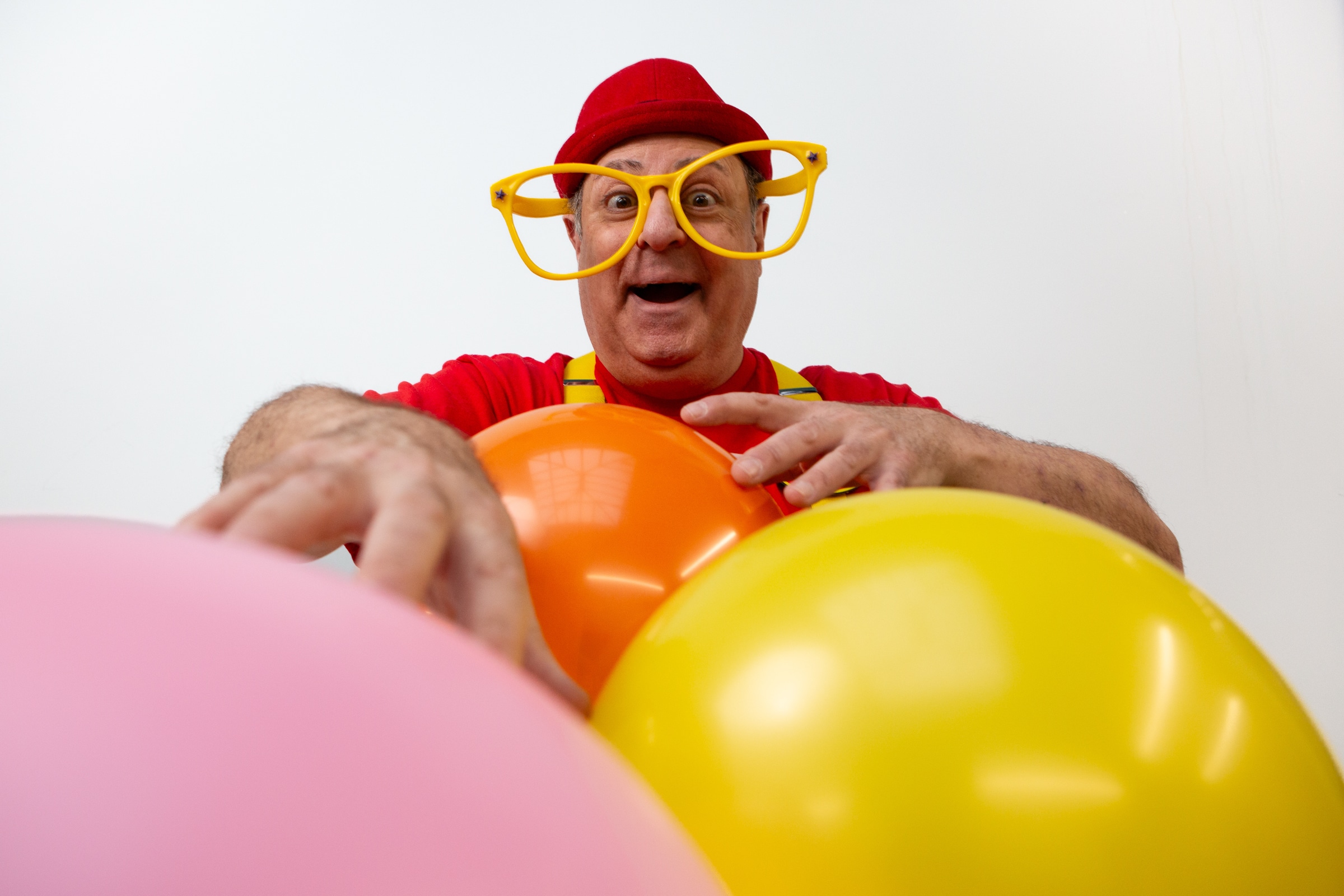 A man with large funny glasses and a red hat holds a pink, orange and yellow balloon.